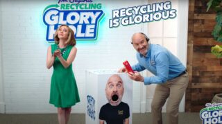 A man and a woman standing next to a box with the words Tom Segura’s Recycling Glory Hole.