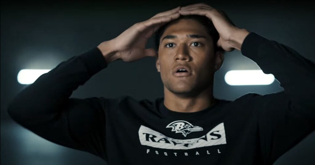 A person with a surprised expression is holding their head with both hands. They are wearing a black shirt with the "Ravens" football team logo. The background is dimly lit, and on the TV behind them, an ad featuring Eli Manning promoting Toyota plays.