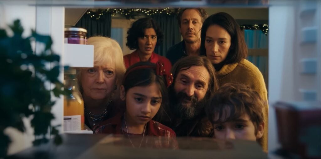 A group of seven people looks intently into an open refrigerator, contemplating the possibilities for the perfect feast in what could easily be a scene from the upcoming Waitrose Christmas Advert 2024.