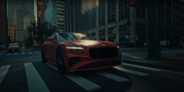 A red Bentley sports car swiftly drives through a city street at dusk. Illuminated skyscrapers provide a stunning backdrop, while a yellow taxi cruises along, reminiscent of a vibrant scene from a Bang Bang Advert.