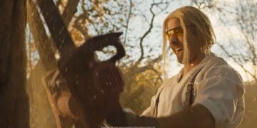 Amidst the greenery, a person with blonde hair and safety glasses expertly wields a chainsaw, as if prepping for the 2025 Ram Big Game Commercial. The scene is reminiscent of Goldilocks and the Three Trucks, where everything is just right in this woodland setting.