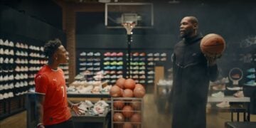 In DICK'S Sporting Goods, two friends discuss the latest buzz as one holds a basketball. Surrounded by athletic shoes and carts of gear, they imagine harnessing IShowSpeed's viral energy for their own game while a hoop stands ready in the background.