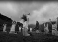In a black and white image, members of the Oat Cult don white robes and plant headdresses in a field. Some kneel while one stands in the center with arms raised. Majestic mountains loom in the background, completing this serene and mysterious tableau.