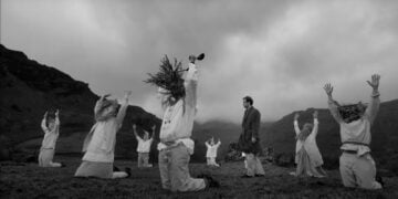 In a black and white image, members of the Oat Cult don white robes and plant headdresses in a field. Some kneel while one stands in the center with arms raised. Majestic mountains loom in the background, completing this serene and mysterious tableau.