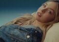 Sydney Sweeney, in a denim jacket for American Eagle, lies on her side with long blonde hair, looking at the camera against a gradient blue background, unfazed by recent ad backlash.