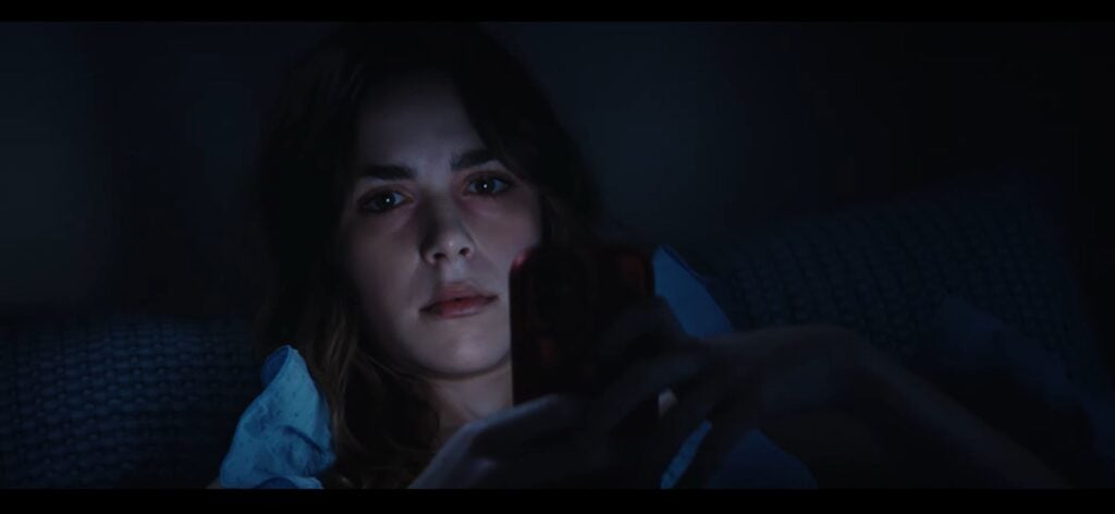A woman lies in bed at night, illuminated by the glow of her smartphone as she scrolls, capturing a scene straight out of a faux horror trailer—Goodnight Phone.