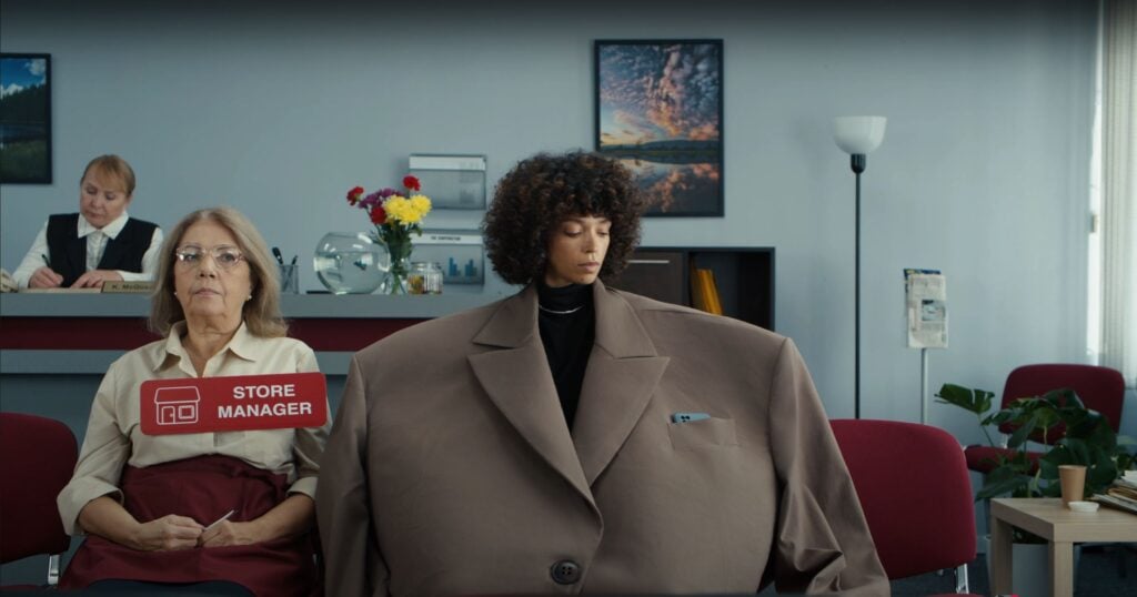 Two women sit in an office waiting area; one wears a "Store Manager" tag, and the other an oversized brown suit. A receptionist works at a desk in the background, possibly conducting a 2025 Career Scout session for Indeed.