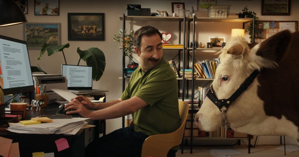 A man at a cluttered desk turns to look at a cow standing beside him in his home office, filled with books and plants—perhaps the most unexpected visitor for his 2025 Career Scout search on Indeed.