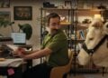 A man at a cluttered desk turns to look at a cow standing beside him in his home office, filled with books and plants—perhaps the most unexpected visitor for his 2025 Career Scout search on Indeed.