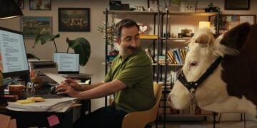 A man at a cluttered desk turns to look at a cow standing beside him in his home office, filled with books and plants—perhaps the most unexpected visitor for his 2025 Career Scout search on Indeed.