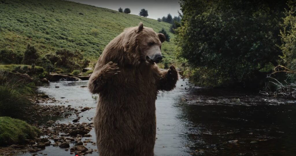 A bear stands upright on its hind legs beside a shallow stream in a green, hilly landscape, evoking the spirit of Nature Valley amid trees and rocks nearby.