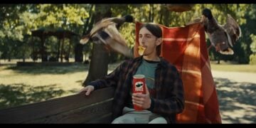 A man with Pringles duck lips sits on a bench, surrounded by flying ducks in a lively park setting.
