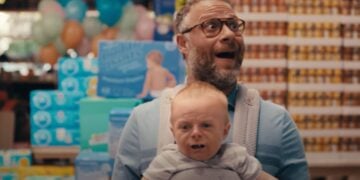 A man with glasses, wearing a baby carrier, stands in a store aisle with a surprised expression while the baby looks upset. Packages of diapers and cans are stacked in the background.