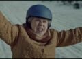 An older woman wearing a blue helmet and a brown quilted coat raises her arms while standing outside on a snowy surface, reminiscent of a joyful moment in an Amazon Holiday Ad.