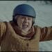 An older woman wearing a blue helmet and a brown quilted coat raises her arms while standing outside on a snowy surface, reminiscent of a joyful moment in an Amazon Holiday Ad.