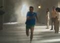 A person in blue and green athletic wear runs toward the camera, showcasing peak performance as debris and dust fill a hallway; five people stand and watch in the background—perfect for an iPhone 17 Pro ad review.