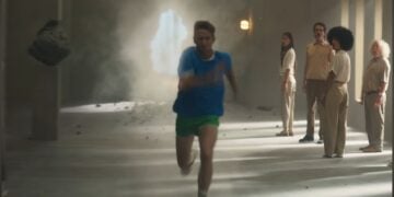 A person in blue and green athletic wear runs toward the camera, showcasing peak performance as debris and dust fill a hallway; five people stand and watch in the background—perfect for an iPhone 17 Pro ad review.