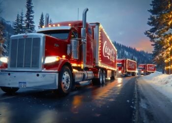 A convoy of red Coca-Cola trucks, featured in an AI-generated ad, glides along a snowy, tree-lined road at dusk, sparkling with festive holiday lights.