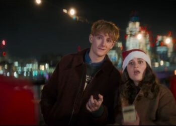 A young man and a young woman wearing a Santa hat stand outdoors at night with city lights in the background, looking toward someone in the foreground—captured with stunning clarity using Pixel 10’s Zoom Power.