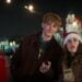 A young man and a young woman wearing a Santa hat stand outdoors at night with city lights in the background, looking toward someone in the foreground—captured with stunning clarity using Pixel 10’s Zoom Power.