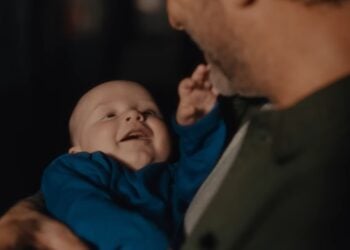 An adult holds a smiling baby dressed in blue, both facing each other in a warmly lit setting—capturing the tender spirit of The Gifting Hour from the 2025 John Lewis Christmas Advert.