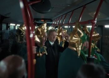 A man in a suit stands on a crowded bus with several people dressed in gold star and green Christmas tree costumes, bringing to life the festive spirit of a Lidl Christmas 2025 ad.