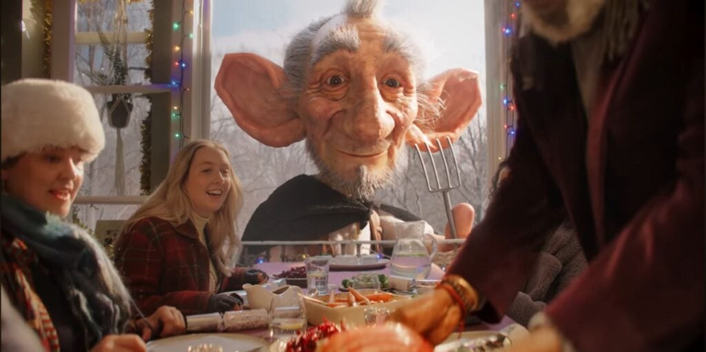 A large, friendly-looking giant peers through a window at a festive table where people are enjoying a meal together, with holiday decorations visible—a magical moment inspired by the Sainsbury’s Christmas 2025 Advert, The Unexpected Guest.