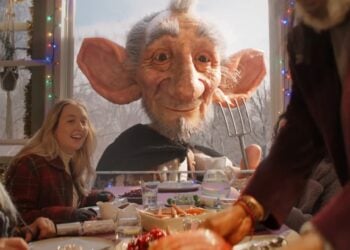A large, friendly-looking giant peers through a window at a festive table where people are enjoying a meal together, with holiday decorations visible—a magical moment inspired by the Sainsbury’s Christmas 2025 Advert, The Unexpected Guest.