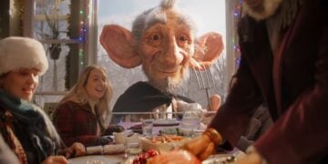 A large, friendly-looking giant peers through a window at a festive table where people are enjoying a meal together, with holiday decorations visible—a magical moment inspired by the Sainsbury’s Christmas 2025 Advert, The Unexpected Guest.