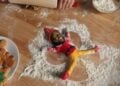 An elf doll with a human face lies on a floured surface in a snow angel pose, surrounded by baking tools and gingerbread cookies.