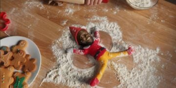 An elf doll with a human face lies on a floured surface in a snow angel pose, surrounded by baking tools and gingerbread cookies.