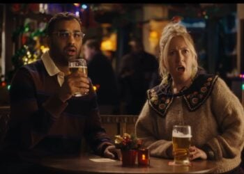 A man and a woman sit at a pub table with drinks, both looking surprised with mouths open. Festive lights twinkle in the background, setting the scene for a charming Mini Rom-Com straight out of a Christmas Ad.