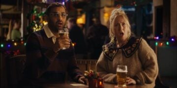 A man and a woman sit at a pub table with drinks, both looking surprised with mouths open. Festive lights twinkle in the background, setting the scene for a charming Mini Rom-Com straight out of a Christmas Ad.