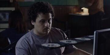 A man with curly hair sits in front of a computer, looking at a tray of several discs—perhaps classic PC Games—extended from the computer in a dimly lit room.