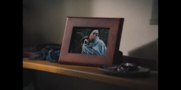 A framed photograph of a person with glasses and a dog sits on a wooden shelf next to a dish holding keys and small items, evoking the cozy feel of a horror-themed ad.