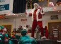 A man dressed as Santa Claus, channeling John Travolta’s Greased Sleigh-Ridin’ style, stands on a workbench surrounded by people in elf costumes inside a festive workshop. Capital One signage is visible in the background.