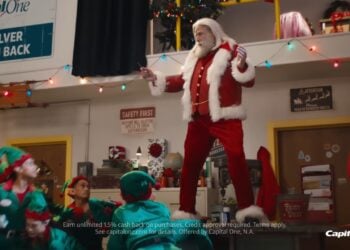 A man dressed as Santa Claus, channeling John Travolta’s Greased Sleigh-Ridin’ style, stands on a workbench surrounded by people in elf costumes inside a festive workshop. Capital One signage is visible in the background.