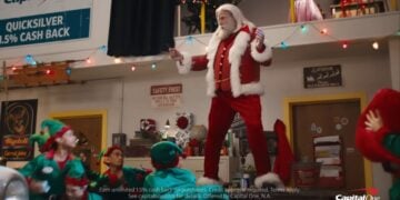 A man dressed as Santa Claus, channeling John Travolta’s Greased Sleigh-Ridin’ style, stands on a workbench surrounded by people in elf costumes inside a festive workshop. Capital One signage is visible in the background.