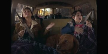 Two people and a dog sit in the backseat of a Chevrolet, covered with blankets. The people appear to be singing or talking with expressive hand gestures, creating a cozy scene straight out of a Memory Lane 2025 Holiday Ad.