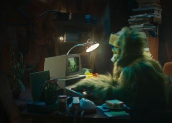 A person in a green furry costume, resembling The Grinch, sits at a cluttered desk using a vintage computer—perfect for a quirky Christmas Ad 2025—with papers, a desk lamp, and office supplies scattered around.