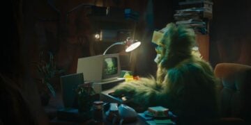 A person in a green furry costume, resembling The Grinch, sits at a cluttered desk using a vintage computer—perfect for a quirky Christmas Ad 2025—with papers, a desk lamp, and office supplies scattered around.
