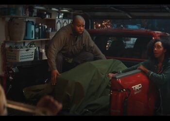 A man kneels in the bed of a red Toyota pickup truck partially covered with a green tarp in a garage, while a woman stands nearby talking to someone off-frame, capturing the cozy spirit of a Toyota Christmas ad 2025.