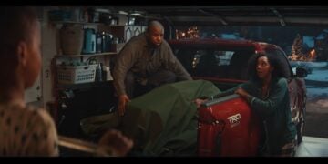 A man kneels in the bed of a red Toyota pickup truck partially covered with a green tarp in a garage, while a woman stands nearby talking to someone off-frame, capturing the cozy spirit of a Toyota Christmas ad 2025.
