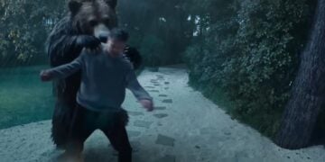 A bear standing on its hind legs appears to attack a person on a stone pathway in a wooded area during heavy rain.