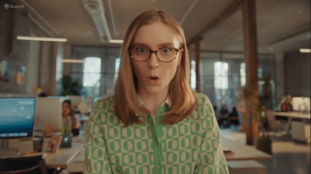 A woman with blonde hair and glasses wearing a green patterned shirt looks surprised in a modern office setting.