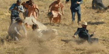 A group of people in formal attire tumble and slide down a dusty hill, with wedding dresses and suits visible.