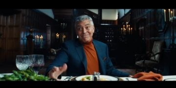 A man in a blue blazer and orange turtleneck sits at a dining table set with food and wine in a dimly lit, elegant room, gesturing with his hand.