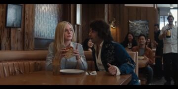 A woman holding a sandwich sits at a diner booth while a man in a blue jacket animatedly talks to her; several people watch and smile in the background.