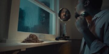 A man shaves in front of a bathroom window at night while a clump of hair with eyes sits on the windowsill, looking at him.