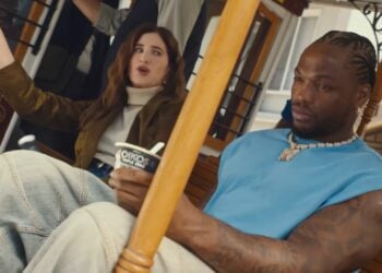 Two people sit side by side on a trolley; the woman gestures animatedly while the man, wearing a blue shirt and chain, holds a cup of Oikos yogurt—perfect for an Oikos Super Bowl 2026 ad review—as he looks ahead.